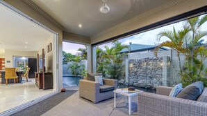 A modern outdoor living area with wicker furniture, a stone wall, and a view of a pool and garden. - Walk to Golf Course, Lagoon Pool, Coastal Lux Home (Pelican Waters)