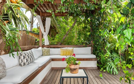 A wooden deck with a white sofa and cushions, surrounded by lush greenery and a pergola.