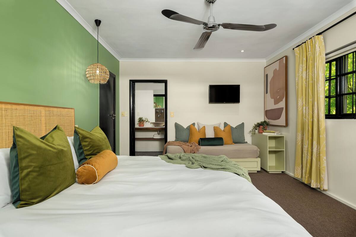 A bedroom with a bed, pillows, a bedside table, a TV, a ceiling fan, and a window with curtains.