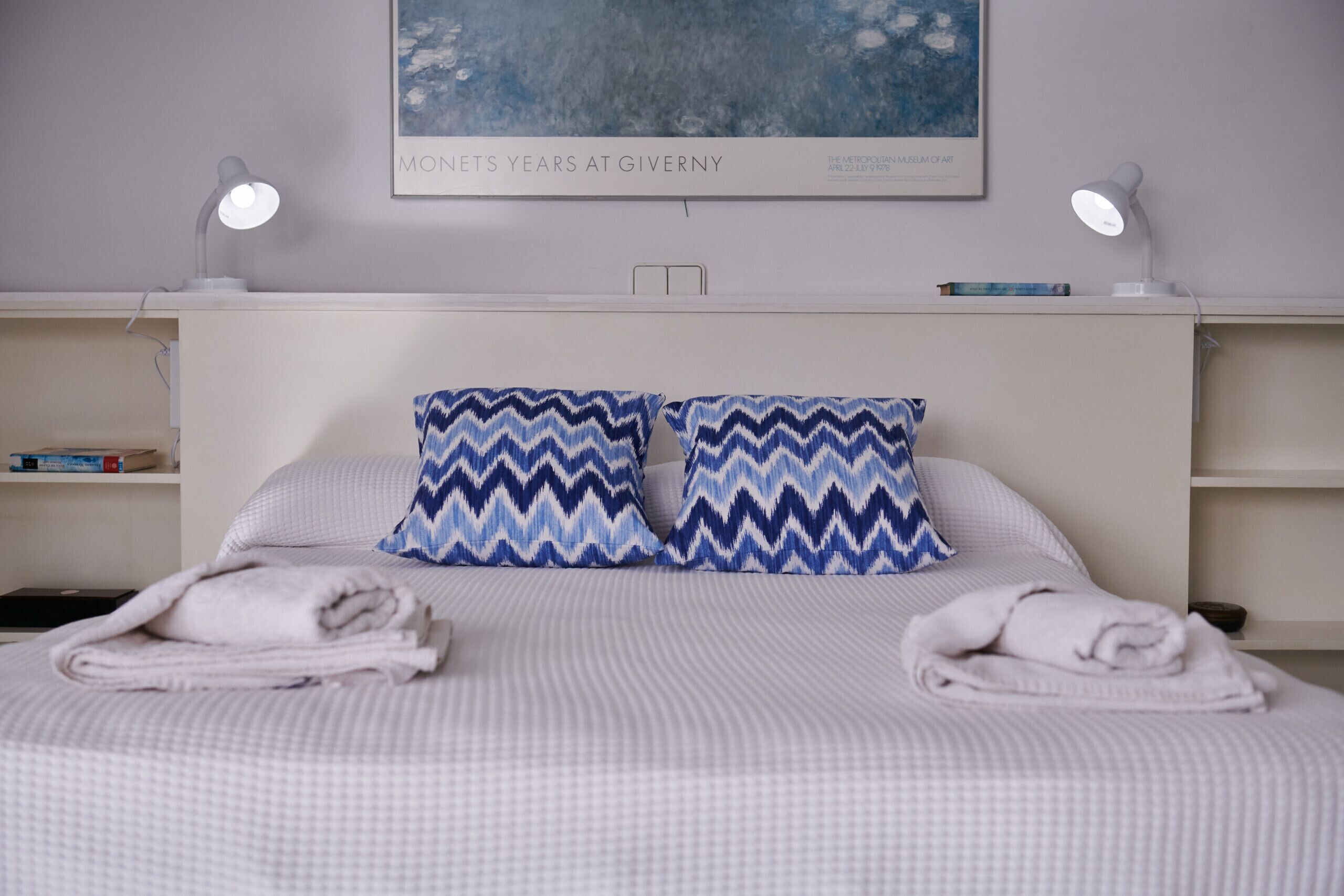 A neatly made bed with two pillows, a bedside table with a lamp, and a shelf with books.