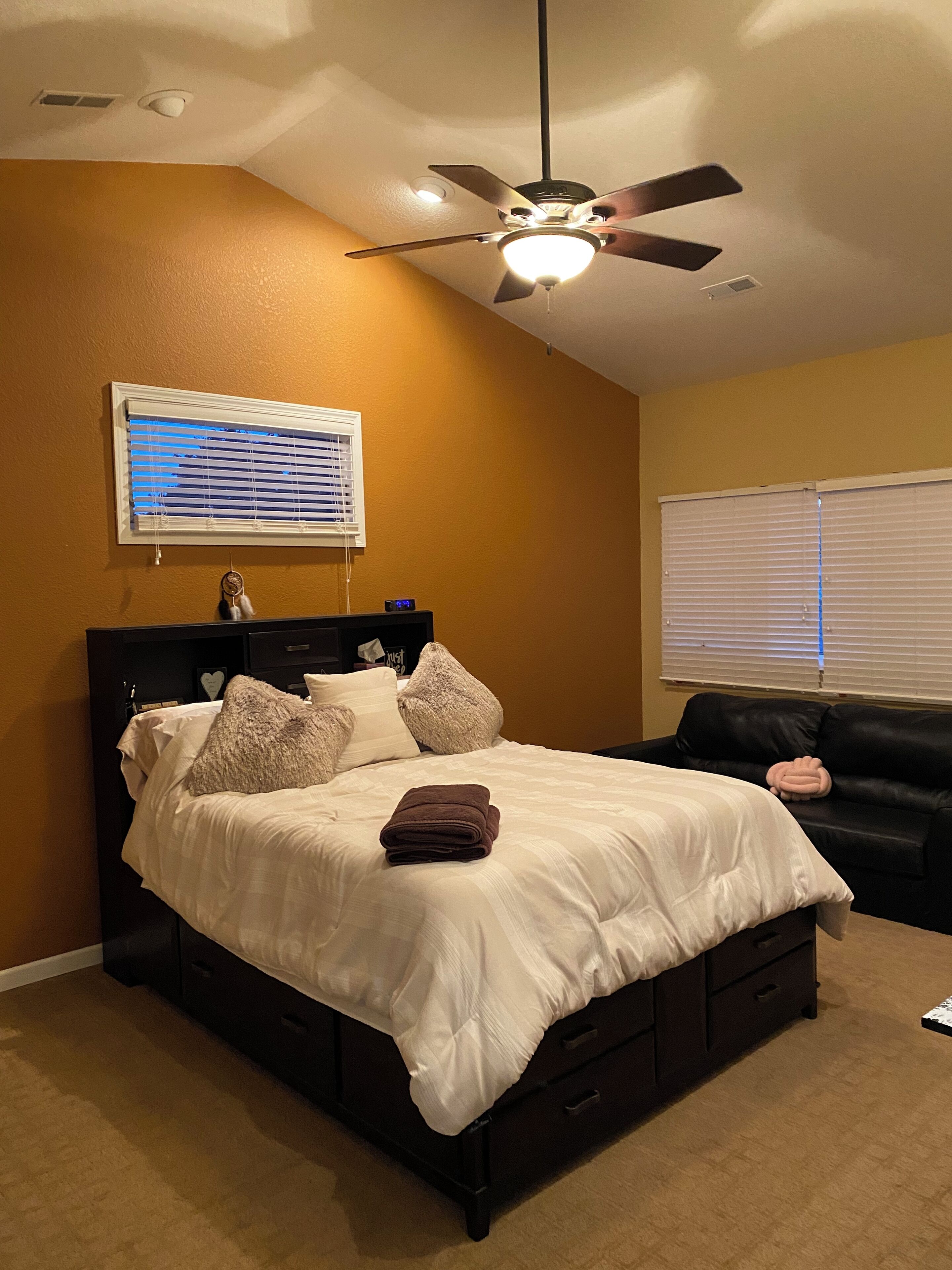 A bedroom with a bed, a ceiling fan, a window with blinds, and a black leather couch.