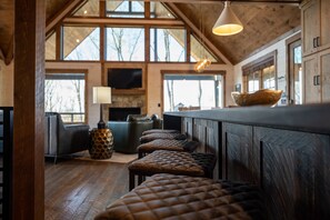 A rustic interior with wooden beams, a stone fireplace, leather ottomans, and a large window overlooking trees.