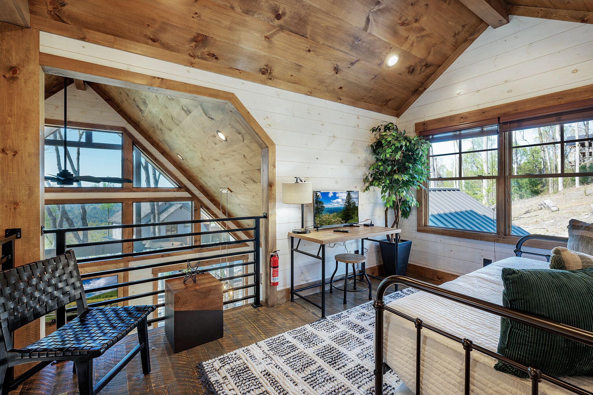 Un acogedor interior de cabaña con techo de madera, un escritorio con computadora, una cama y ventanas amplias que ofrecen vistas del exterior.