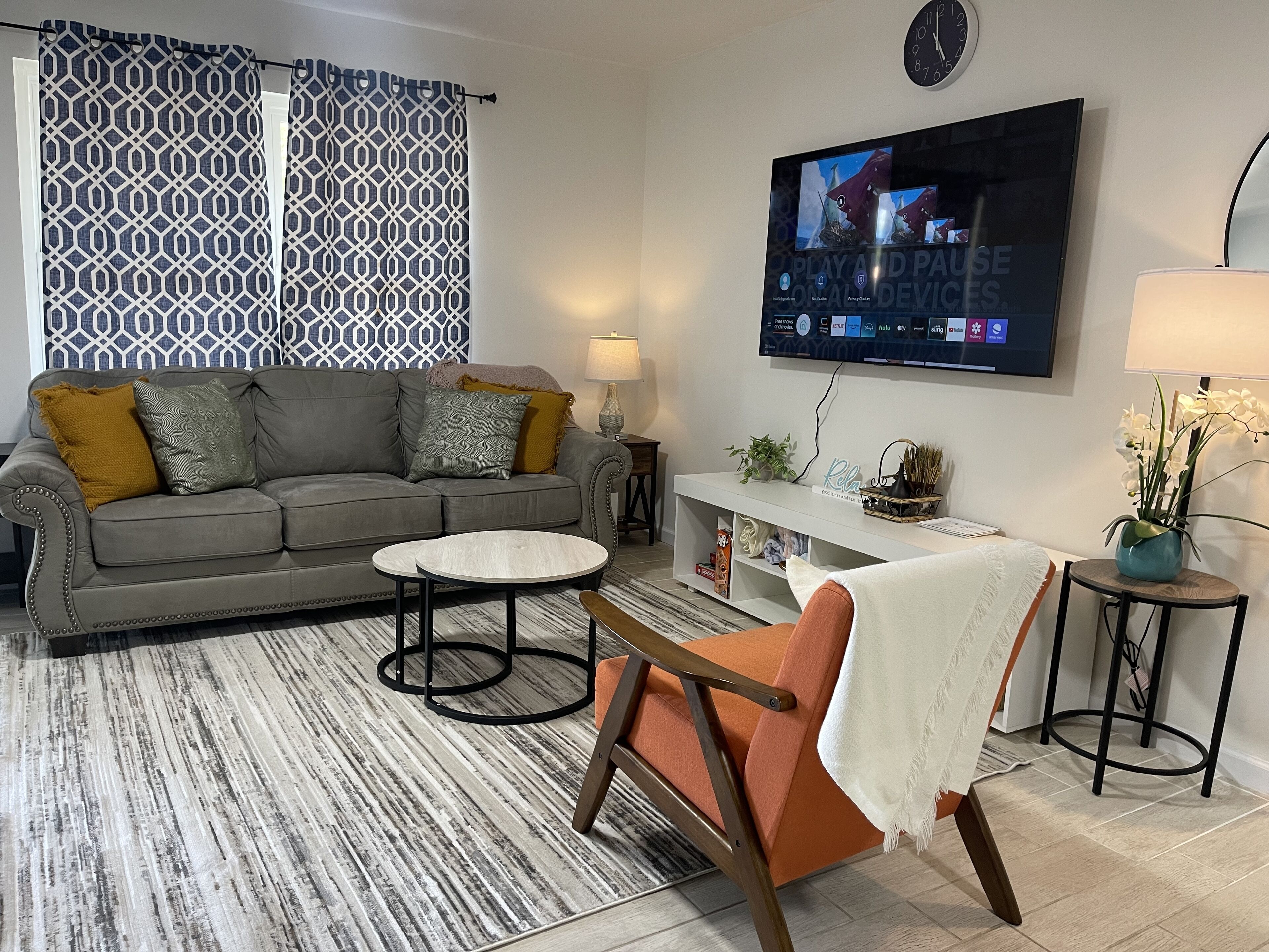 A living room with a grey sofa, a round coffee table, an orange armchair, a TV mounted on the wall, and a patterned curtain.