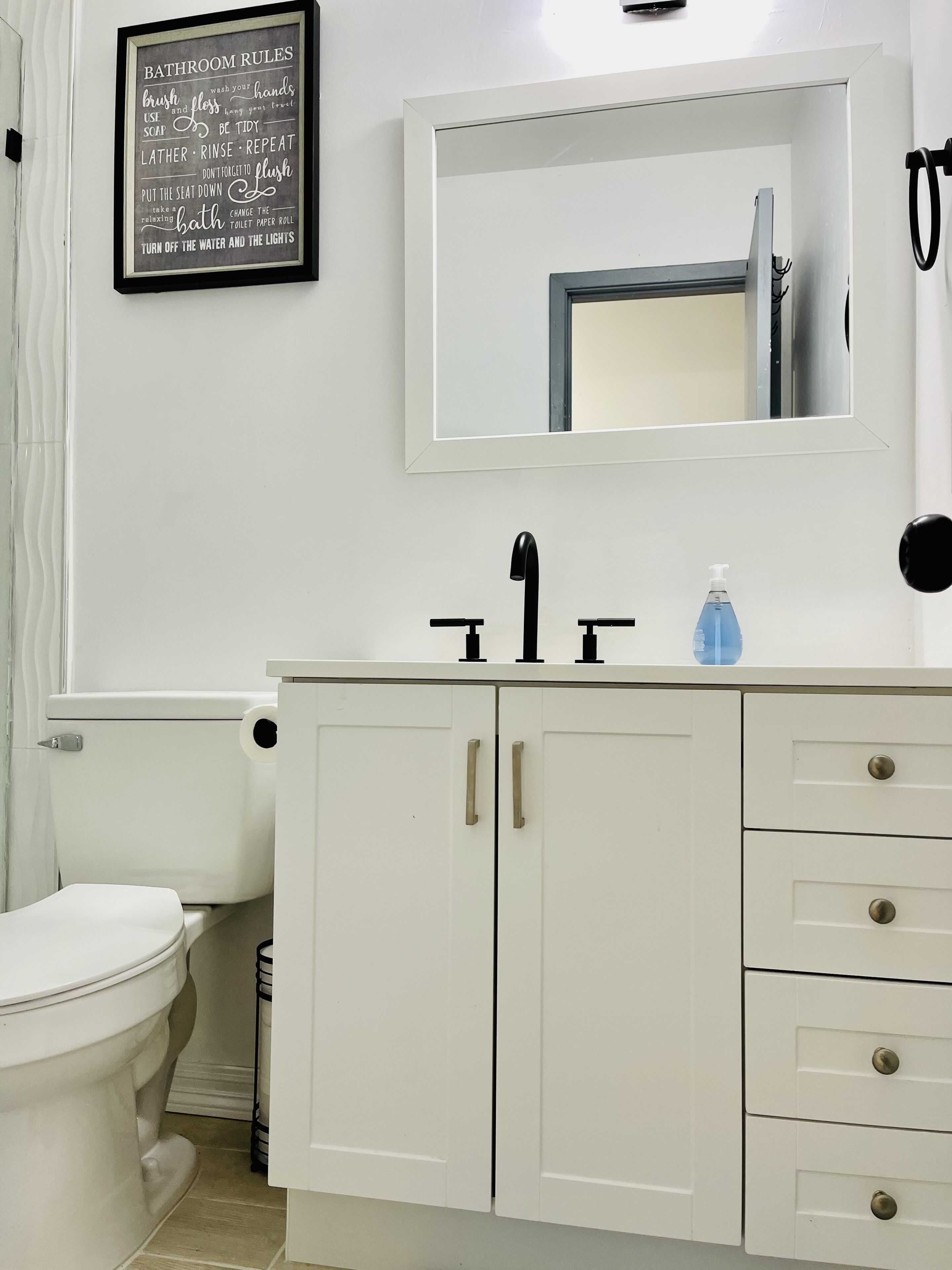 Une salle de bain comprenant des toilettes, un lavabo avec un miroir au-dessus, et un panneau encadré affichant les règles d’utilisation de la salle de bain.