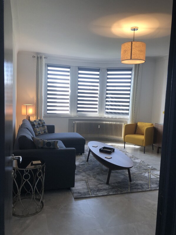 Un salon moderne avec un canapé, une table basse et un fauteuil jaune. De grandes fenêtres sont équipées de stores, il y a un radiateur et une lampe suspendue.