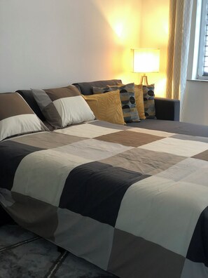 Una cama bien hecha con una colcha a cuadros, una lámpara de noche y una ventana con cortinas.