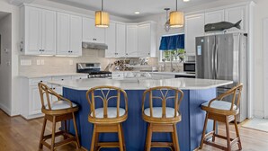 Une cuisine avec des meubles blancs, un grand îlot central doté de tabourets, des appareils en acier inoxydable et des luminaires suspendus.