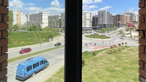 A view from a window with brick framing, overlooking a cityscape with multiple buildings, a road with vehicles, and a grassy area. - SunnyHill Apartments (Pristina)