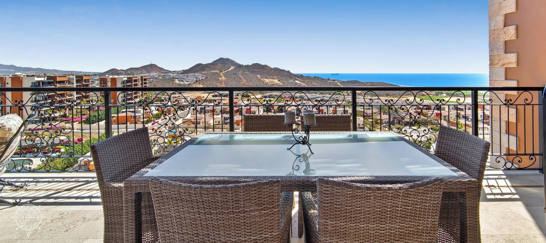 Un balcón con sillas de mimbre y una mesa de vidrio, con vistas a un pueblo costero y montañas a lo lejos.