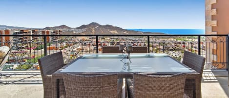 A balcony with wicker chairs and a glass table, overlooking a coastal town with buildings and mountains in the distance.