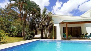 A swimming pool with a covered patio area, surrounded by lush greenery and palm trees.