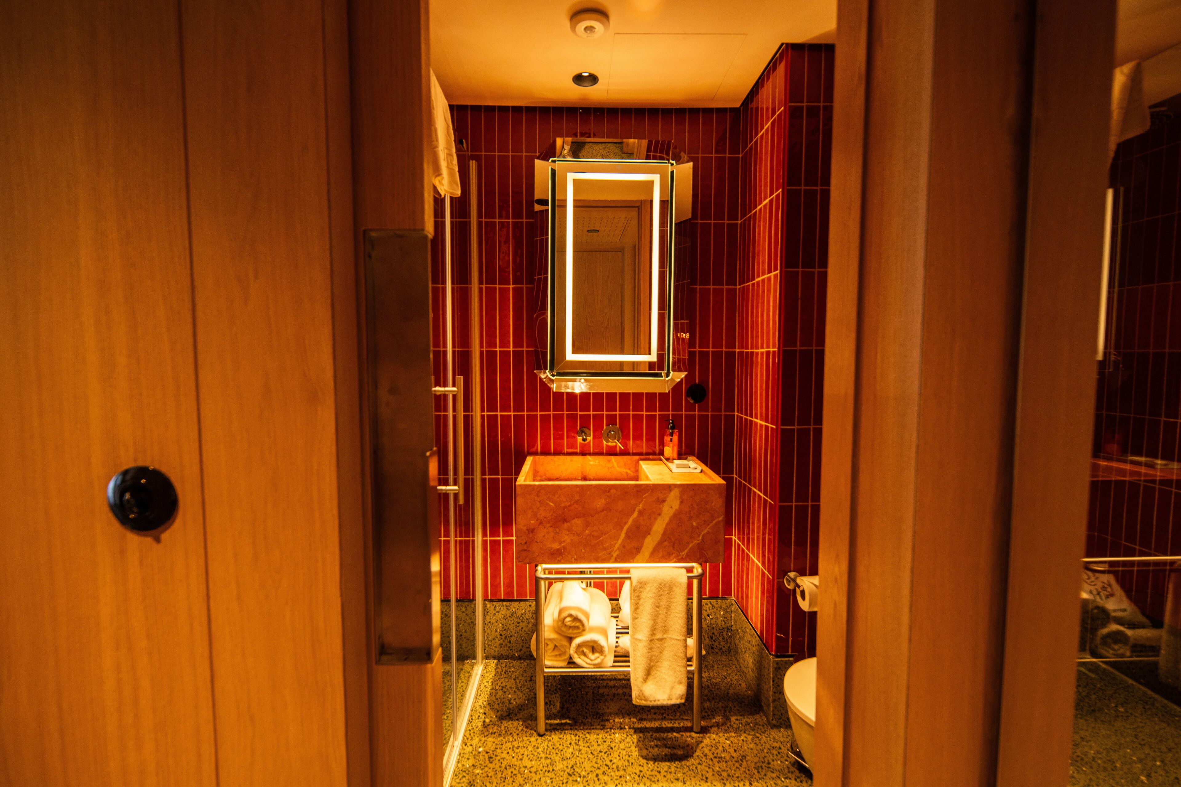 A narrow hotel bathroom with a wooden door, a sink with a mirror, and a towel rack.