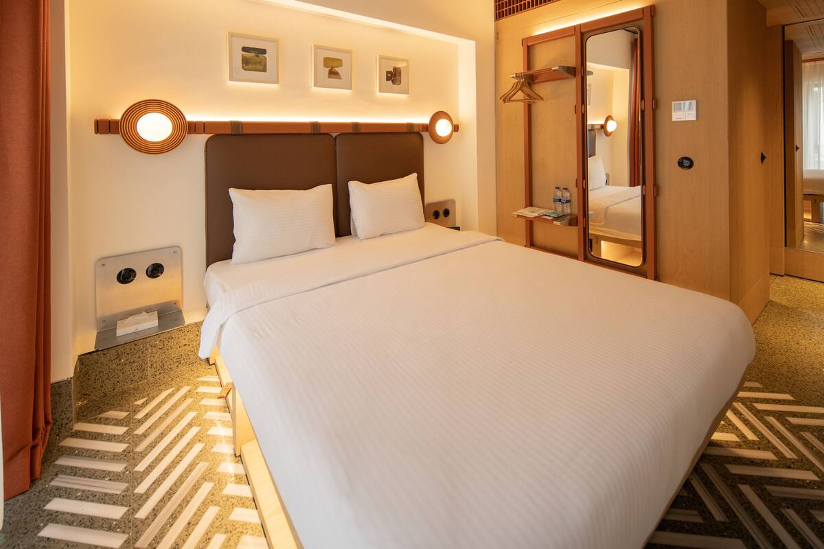 A hotel room with a large bed, a wardrobe, a mirror, and a wall-mounted light fixture.