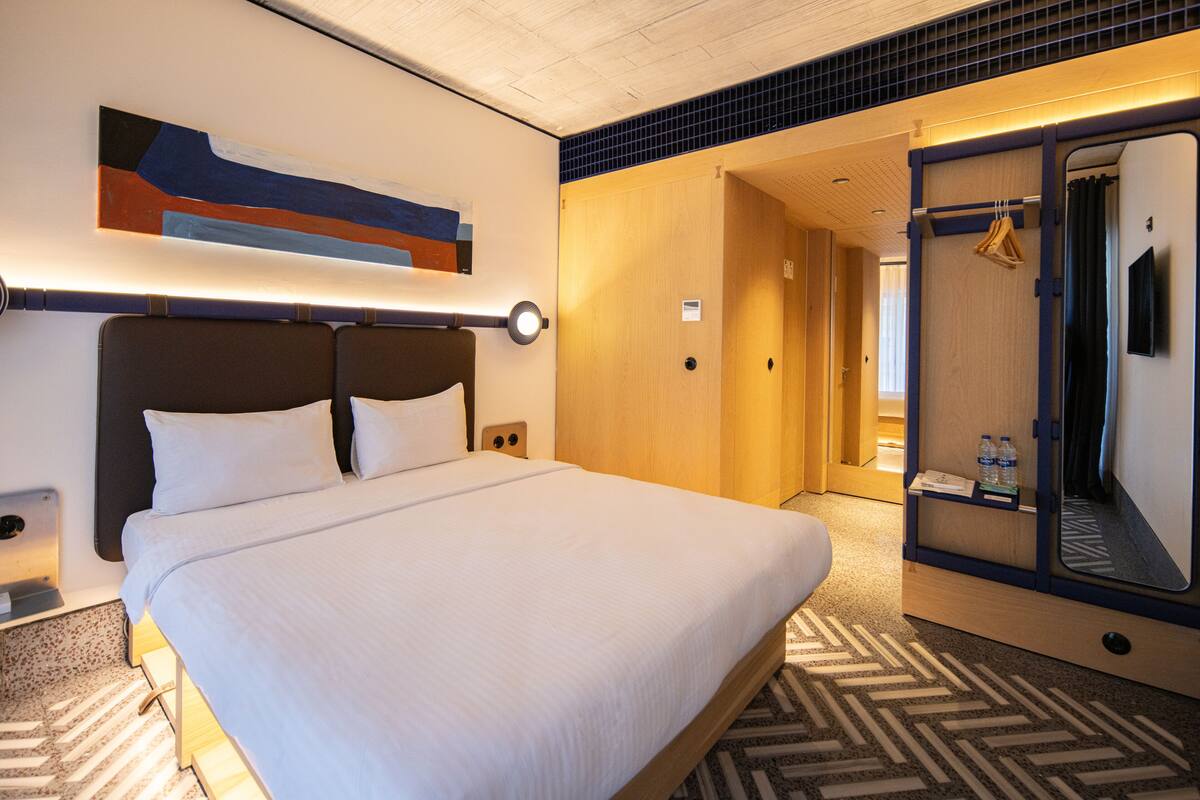 A hotel room with a large bed, a headboard, a wall-mounted artwork, a mirror, and a door leading to a bathroom.