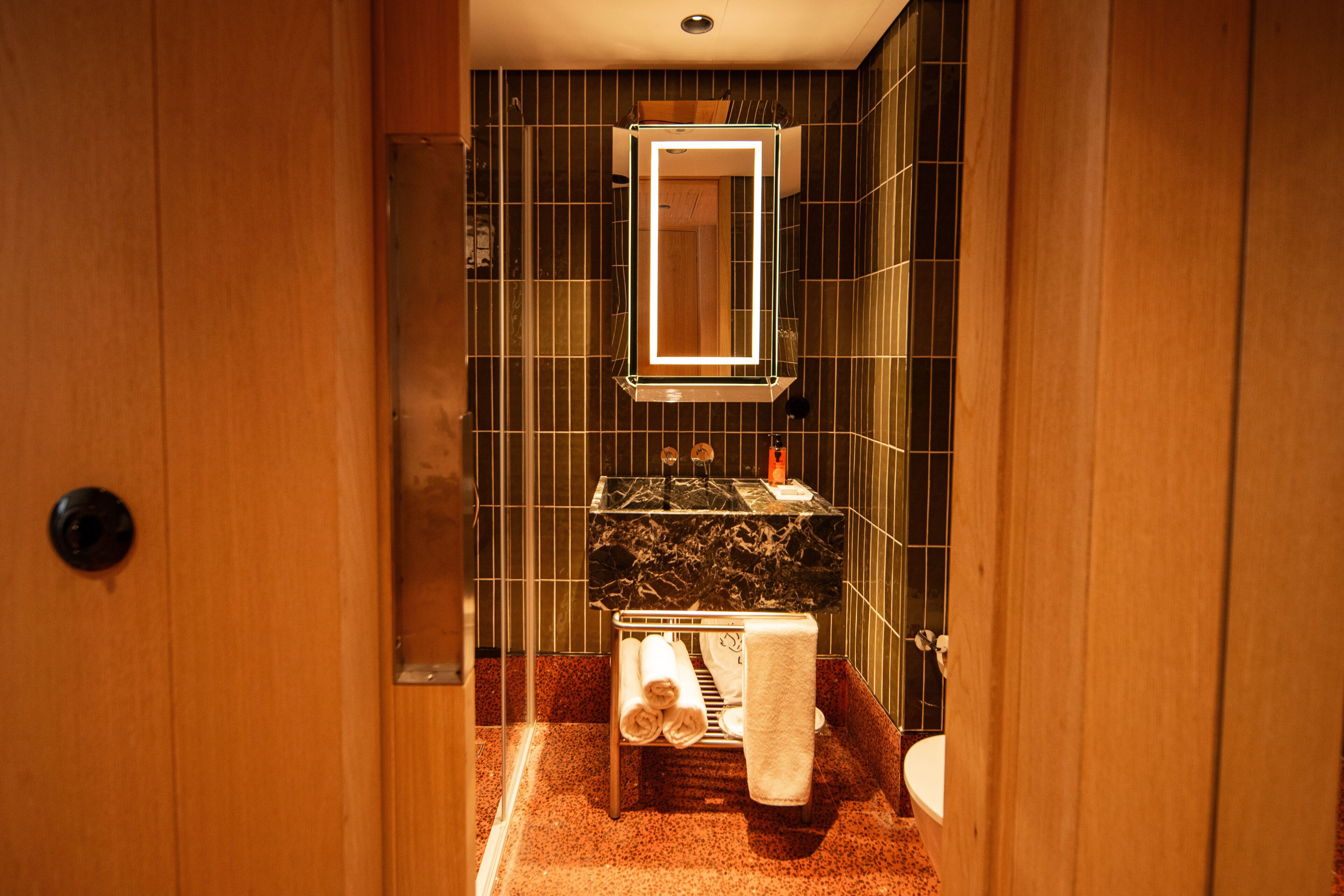 A narrow bathroom with a wooden door, a mirror with illuminated frame, a sink with a black countertop, and a towel rack with folded towels.