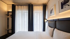Una habitación de hotel con una cama grande, una mesita de noche, un perchero y una ventana con cortinas.