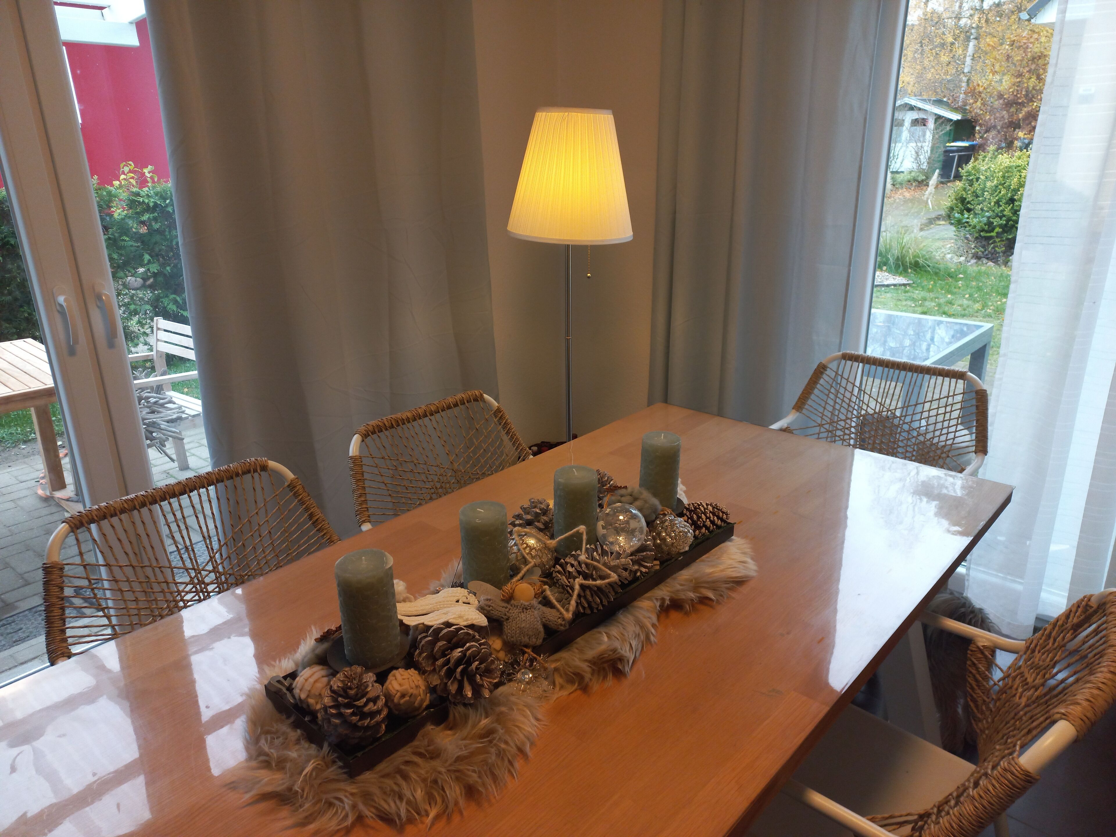 Una mesa de comedor con velas, piñas y un mantel de piel. Hay sillas de mimbre alrededor de la mesa. Una lámpara está sobre la mesa, y se ve una ventana con cortinas al fondo.