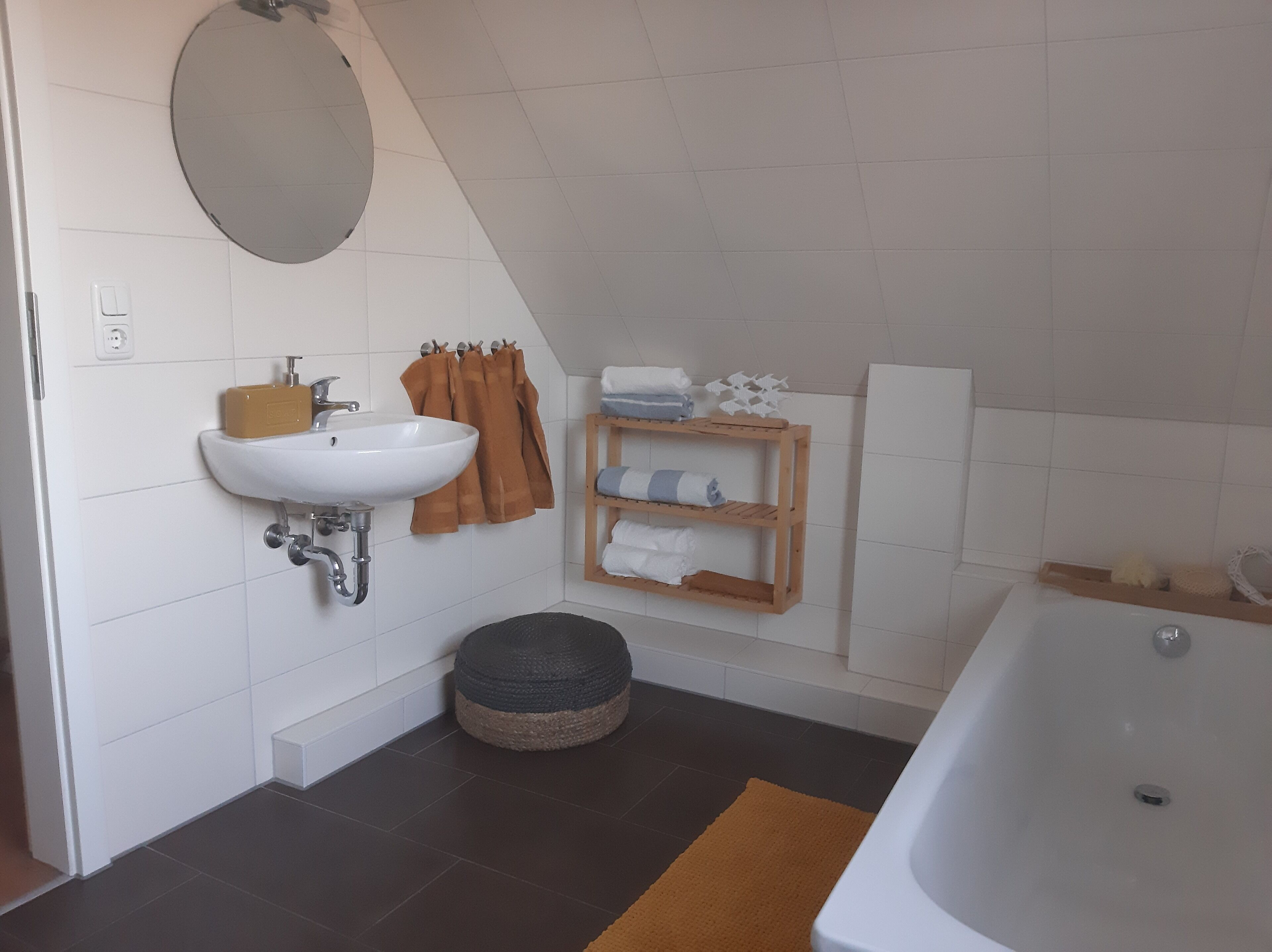 A bathroom with a white sink, a round mirror, a wooden shelf with towels, a round ottoman, and a bathtub.