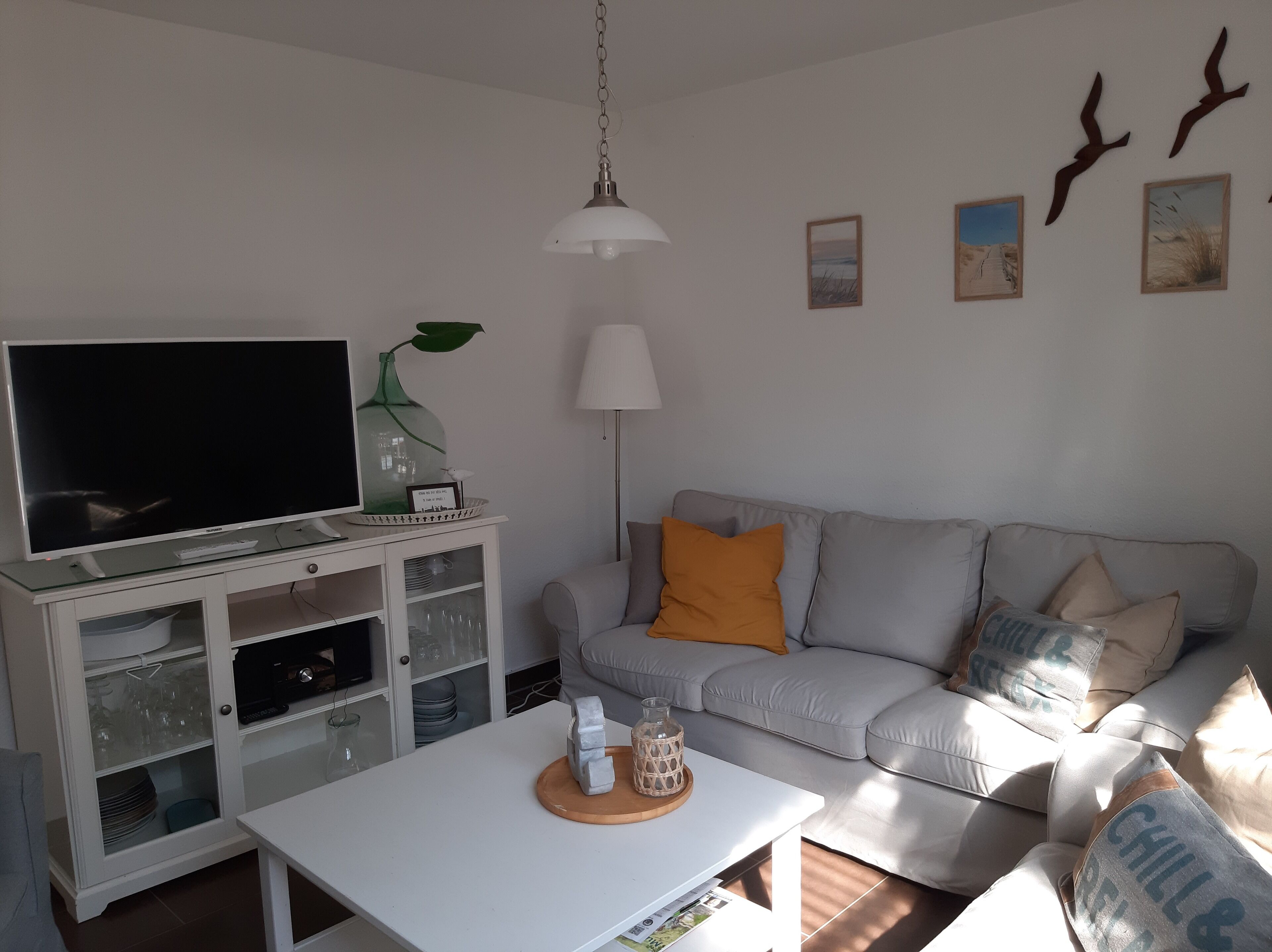 A living room with a television, a white cabinet, a grey sofa with orange cushions, a white table, and a hanging lamp.