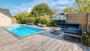A wooden deck with a swimming pool, outdoor seating, and a view of a house in the background. - Villa with heated pool & garden - 5 min from the sea, by Groom (Guidel)