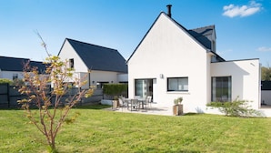 Une maison moderne à deux étages, avec une façade blanche, un toit foncé et un petit jardin comprenant une pelouse et un arbre.