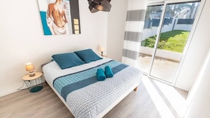 Une chambre moderne avec un lit, des oreillers, une table de chevet, une lampe et une grande fenêtre donnant sur un jardin.