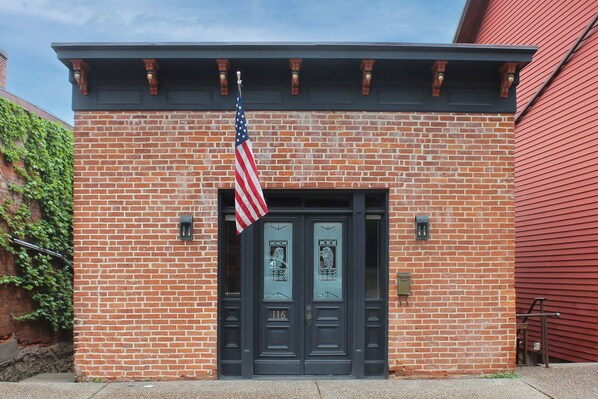 A brick building with a dark-colored entrance door, an American flag, and a number "116" on the door.