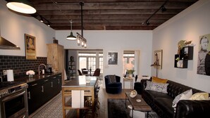 A modern kitchen with a central island, dark wooden cabinets, and a black countertop. There is a dining area with a black leather sofa and a coffee table. The room has a rustic ceiling with exposed beams and pendant lighting. - The Owl House, 1850's Historic Home: Luxury Steps from Downtown  (Galena)