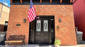 A brick building with a black door, an American flag, and a bench outside. - The Owl House, 1850's Historic Home: Luxury Steps from Downtown  (Galena)