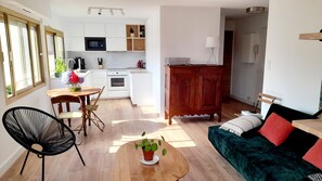 Cocina moderna con armarios blancos, microondas y cafetera. Hay un comedor con mesa y sillas de madera, y un sofá con cojines.
