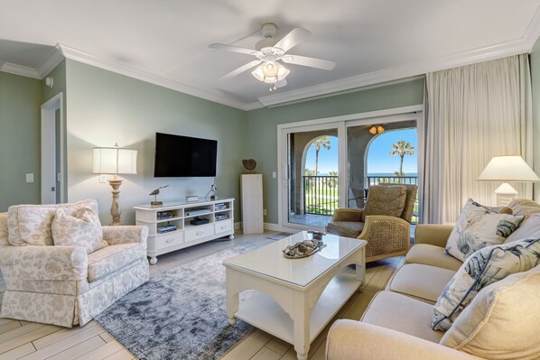 Una sala de estar con ventilador de techo, televisor de pantalla plana, mueble blanco para equipo multimedia y vistas al exterior.