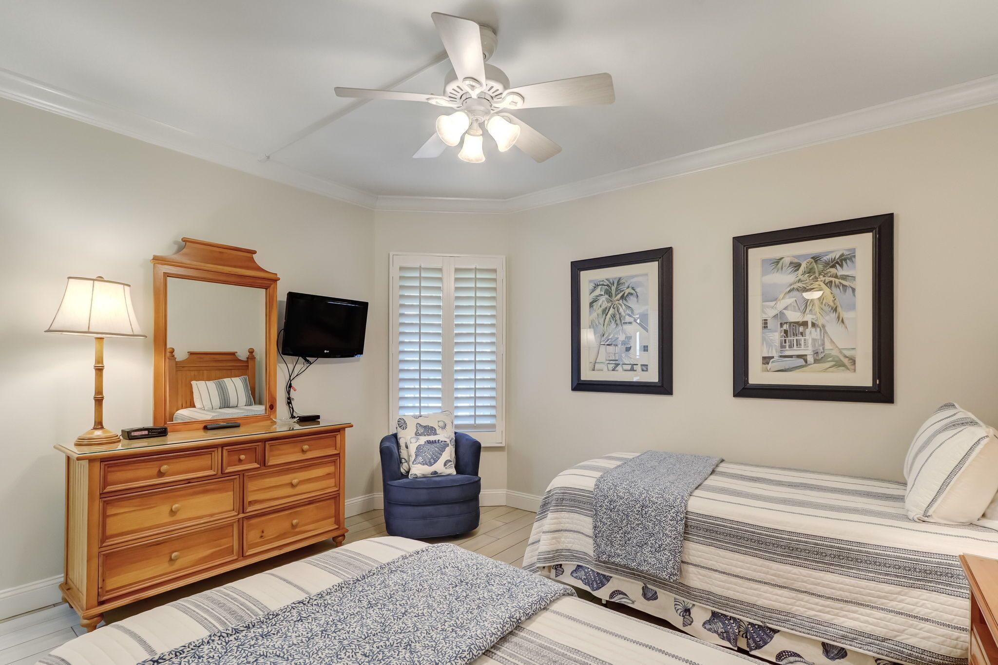 A bedroom with a wooden dresser, a chair, two beds, a ceiling fan, a TV, and two framed pictures on the wall.