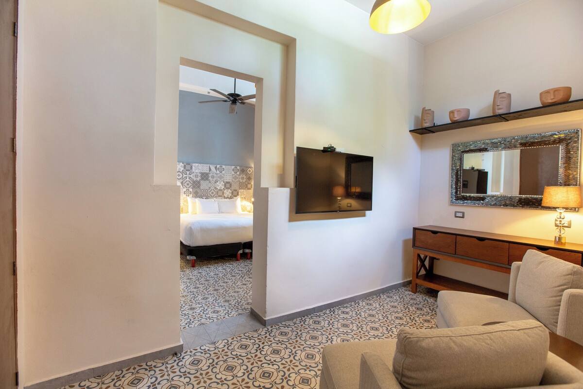 A hotel room with a bed, a TV, a mirror, a sofa, and patterned floor tiles.
