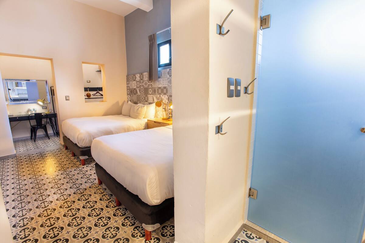 A hotel room with two beds, a desk with a chair, a mirror, and a patterned floor.
