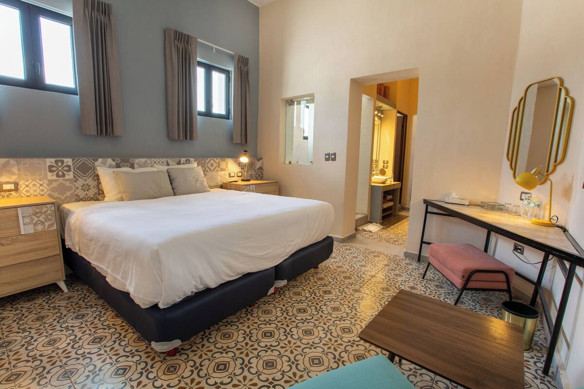 A bedroom with a large bed, bedside tables, a desk, a chair, and a small stool.