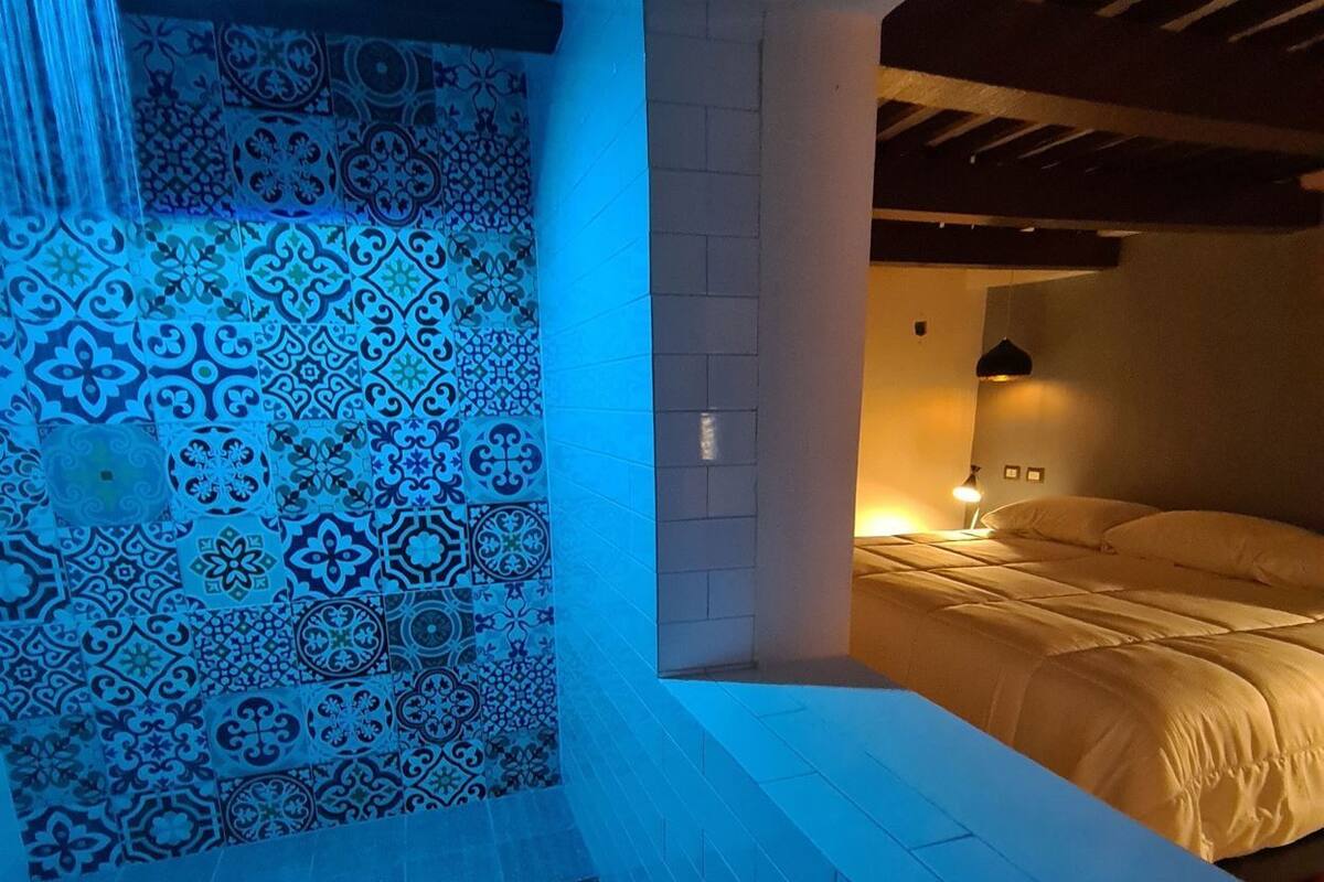 A room with a patterned tile wall, a bed, a small table, and a wall-mounted lamp.