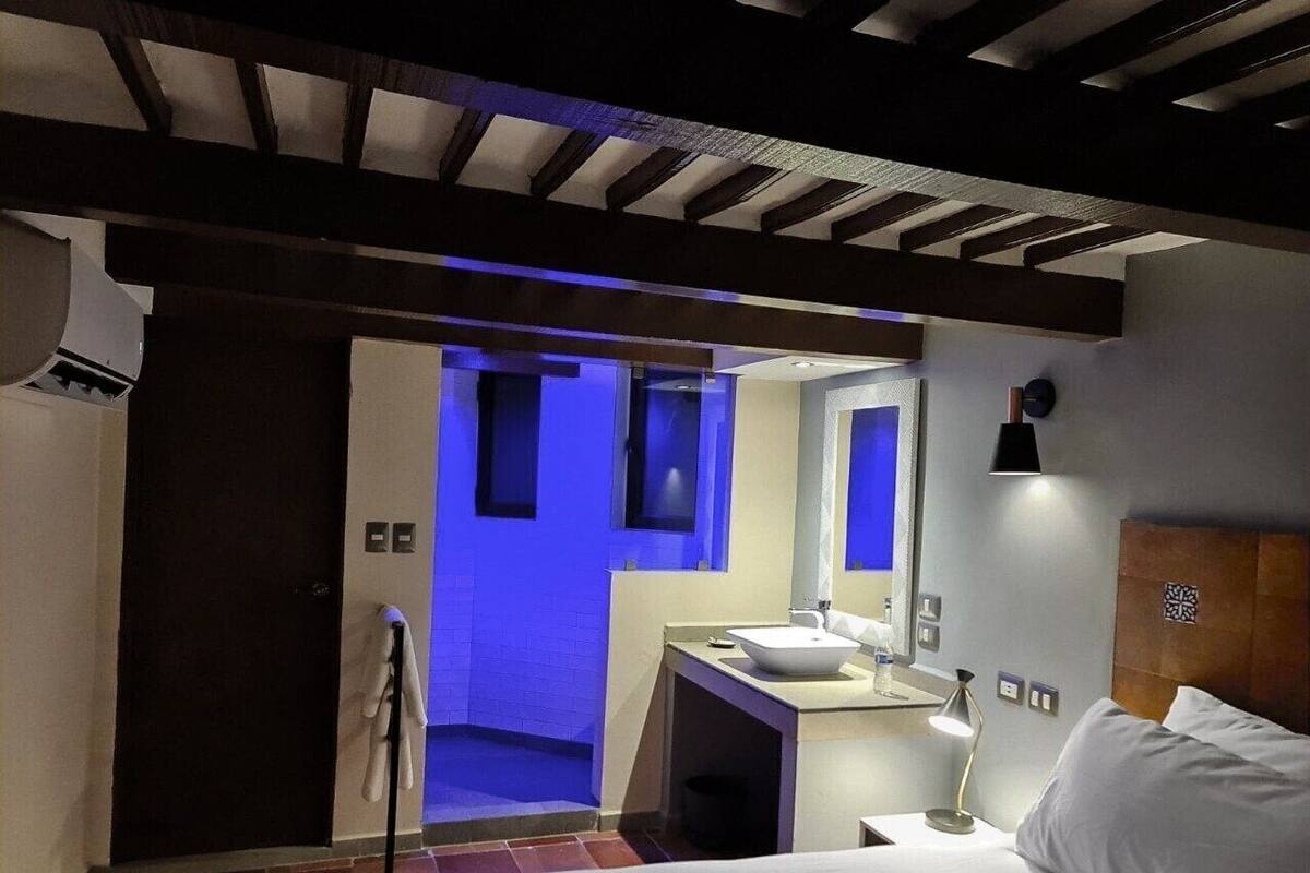 A modern hotel room with a bed, a bedside table, a nightstand, a large mirror, and a bathroom visible through an open door.