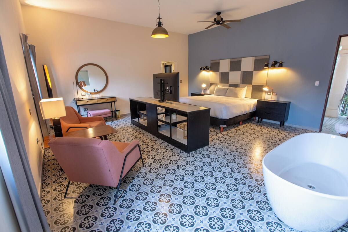 A modern hotel room with a checkered floor, a large bed, a desk, a chair, a TV, a ceiling fan, and a bathtub.