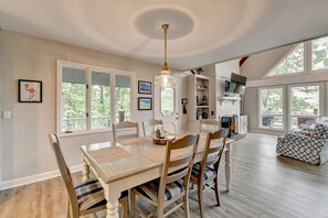 A dining area with a wooden table and chairs, a chandelier, and a view of the outdoors through large windows.