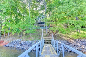 A wooden bridge leading to a house surrounded by dense green trees.