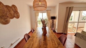 A dining room with a wooden table, cushioned chairs, a woven light fixture, and a view through large windows. - AP376 VALPARAISO (Nerja)