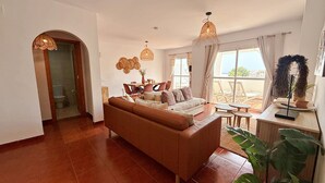A living room with a brown sofa, a dining table with chairs, a large window with curtains, and a balcony with plants. - AP376 VALPARAISO (Nerja)