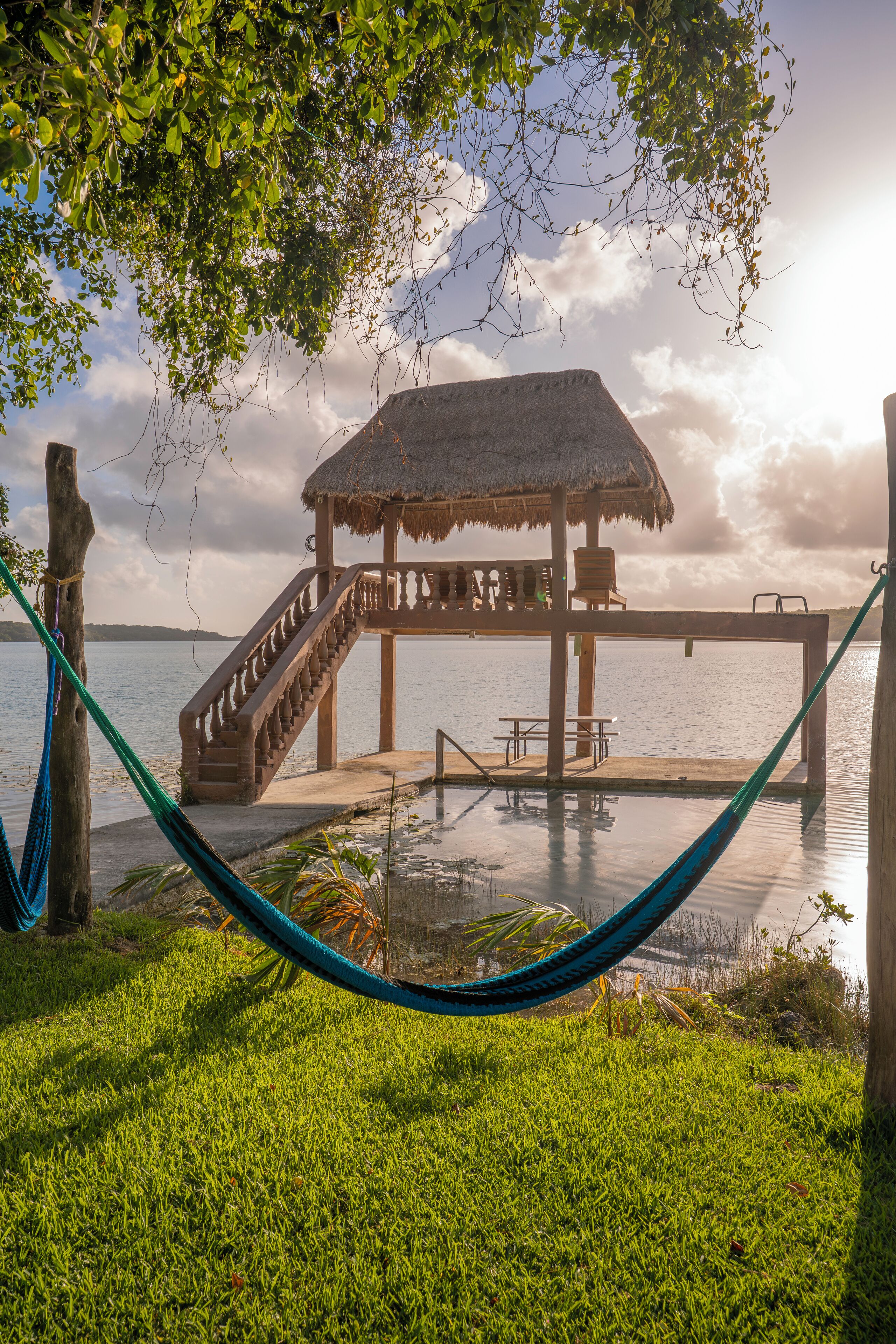 A wooden dock with a thatched roof, a staircase leading to a gazebo, and a hammock in the foreground.