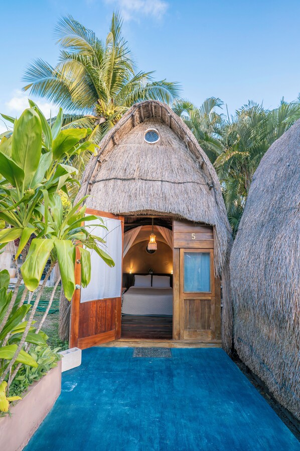 Estructura de techo de paja con puerta de madera y un cartel con el número '5', rodeada de plantas tropicales y una piscina azul.