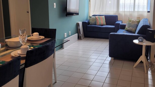A modern living room with a flat-screen TV, a blue sofa, and a dining area with a table set for two. - Apartamento da Cor do Mar - VR-1202 (Recife)