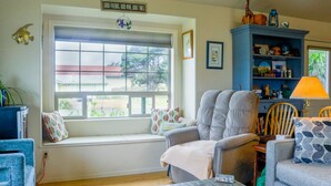 Ein gemütliches Wohnzimmer mit großem Fenster, einem bequemen Sessel, einem blauen Bücherregal und einem kleinen Tisch.
