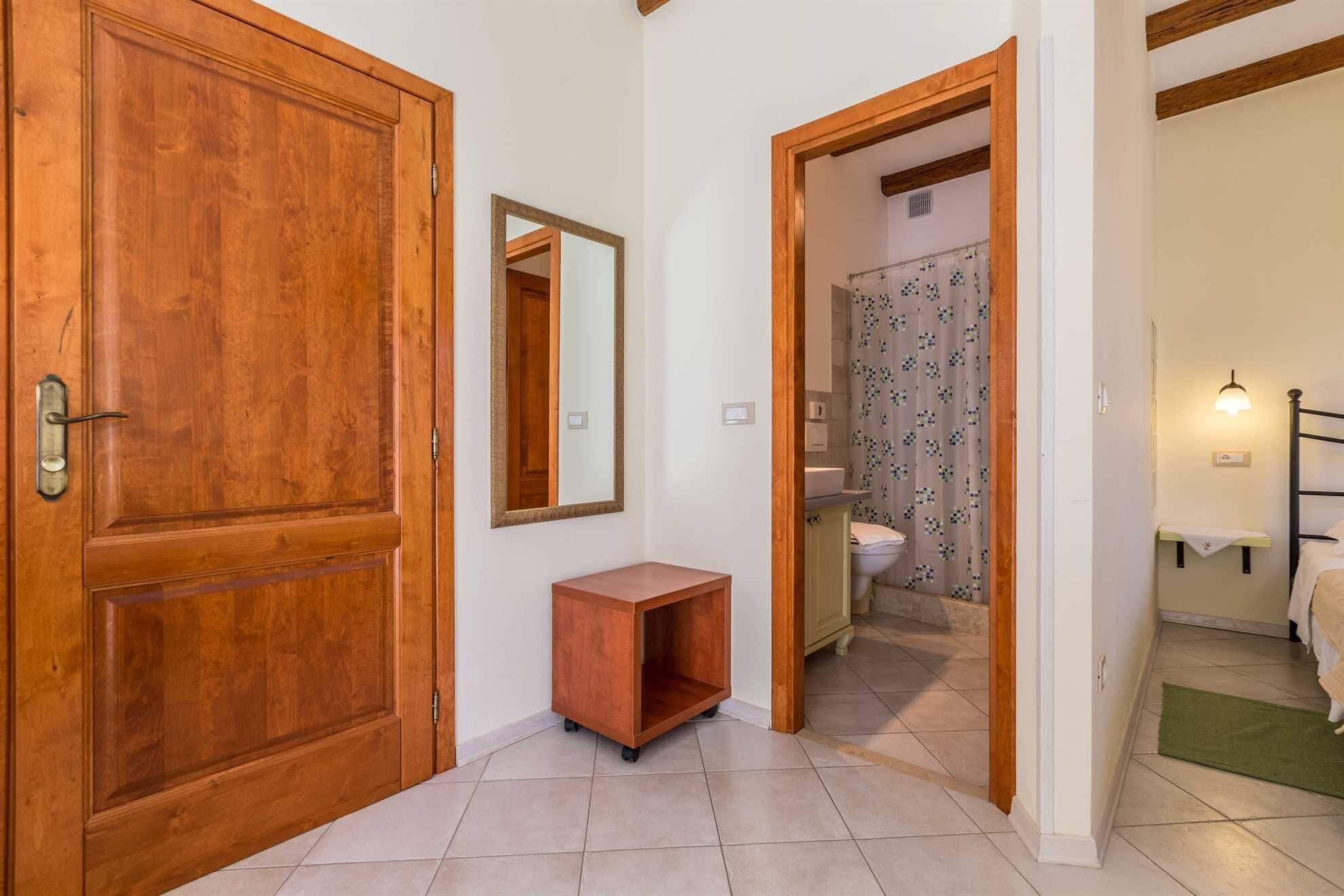 Un couloir avec une porte en bois, un miroir, une petite armoire en bois et une salle de bain visible à travers une porte ouverte.