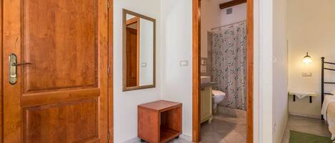 Un couloir avec une porte en bois, un miroir, une petite armoire en bois et une salle de bain visible à travers une porte ouverte.