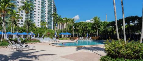 A hotel pool area with lounge chairs, palm trees, and a large building in the background.